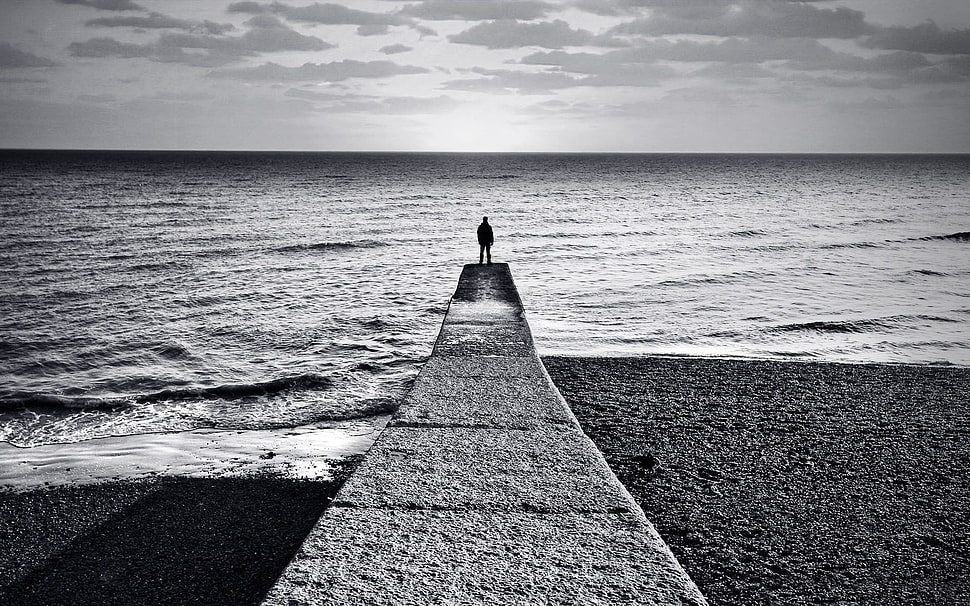 grayscale photo of man standing on bridge beside beach shore HD wallpaper