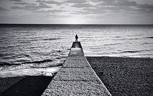 grayscale photo of man standing on bridge beside beach shore