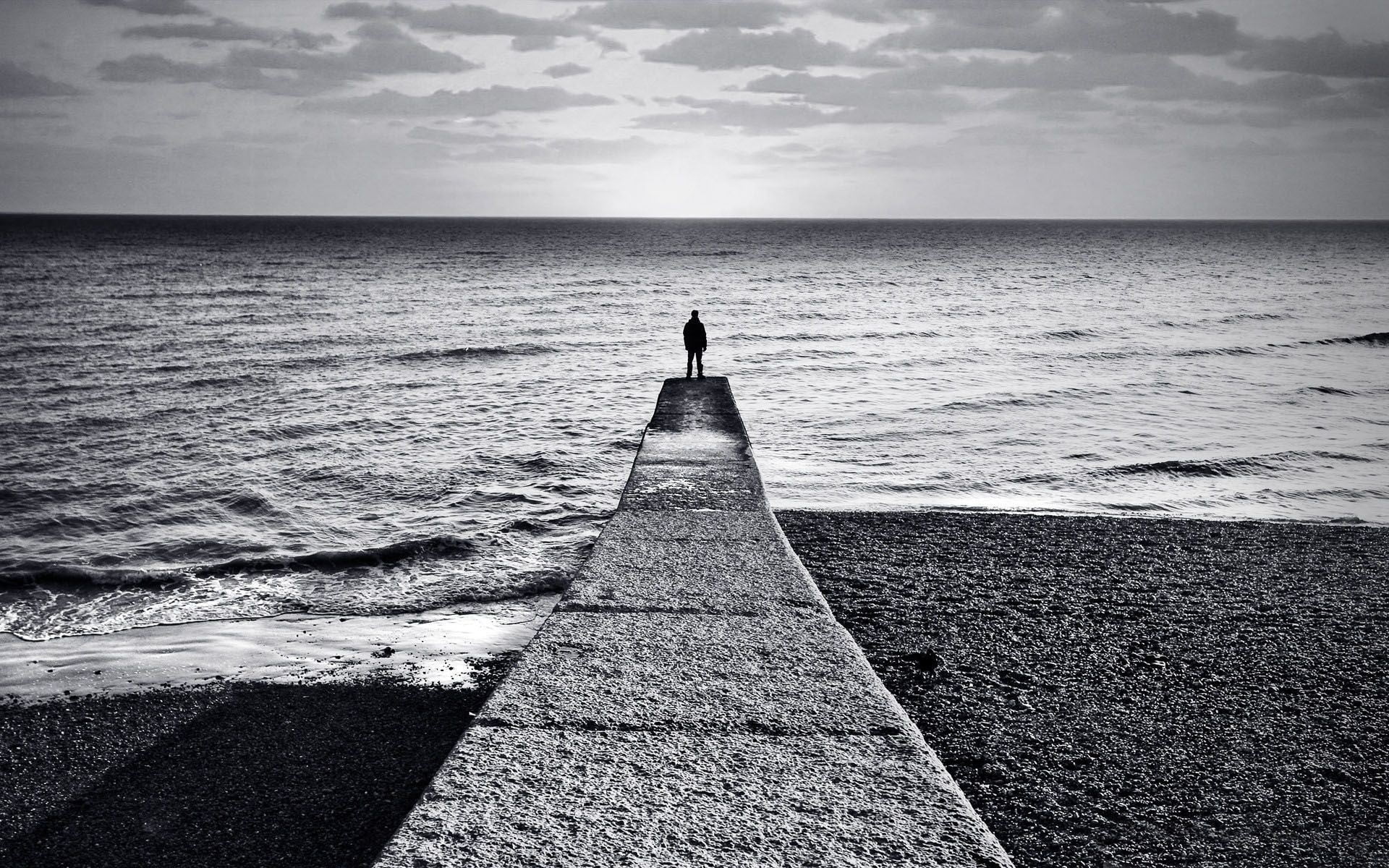 grayscale photo of man standing on bridge beside beach shore