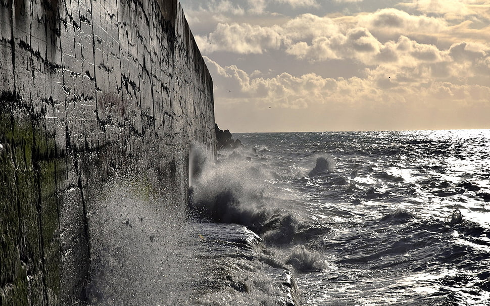 gray concrete wall beside ocean waves under cloudy sky during day time HD wallpaper