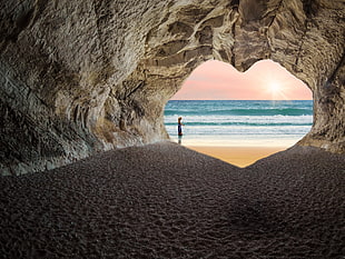 photo of person walking on seashore in front of cave during day time