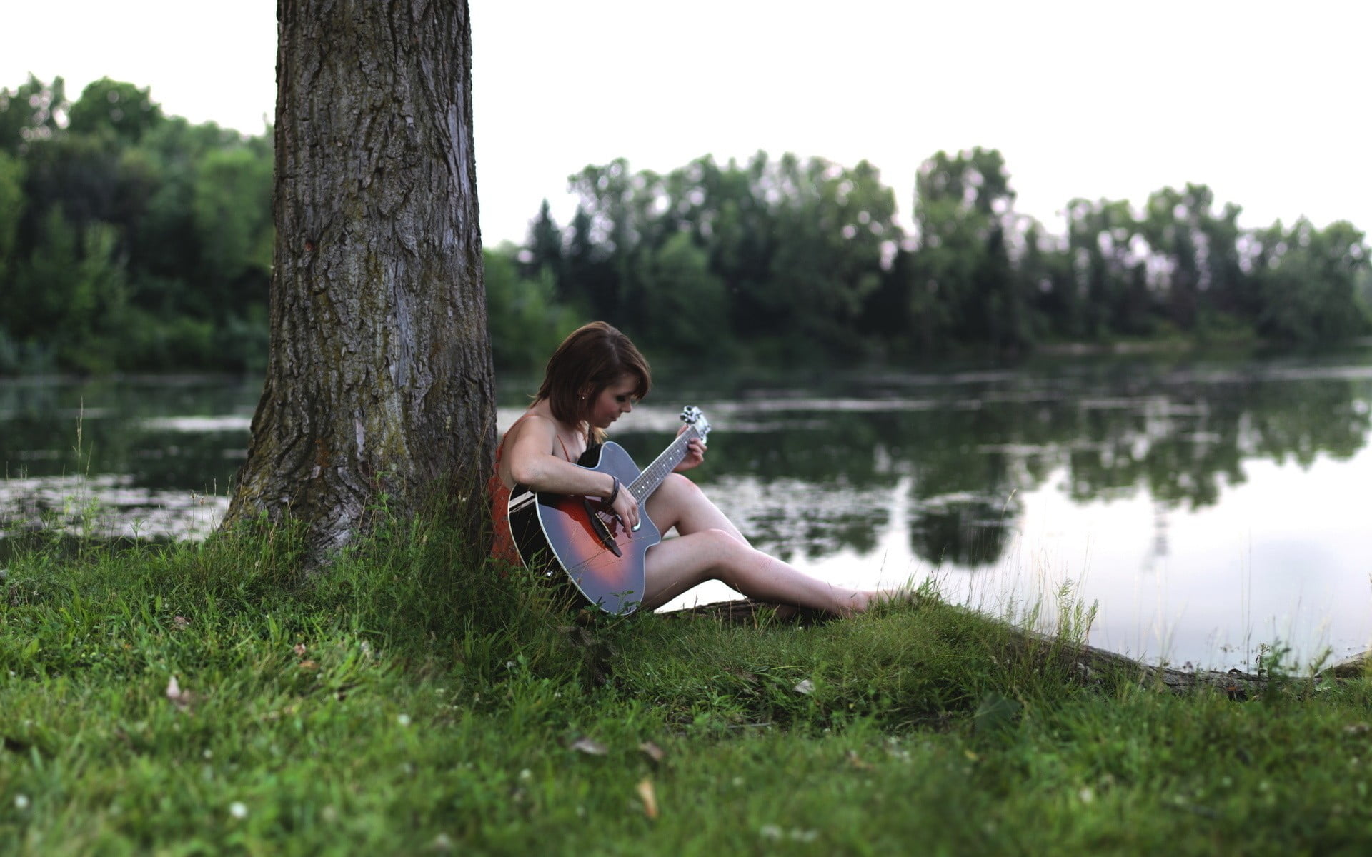 selective focus photo of a woman sitting at the back of the tree about to play acoustic guitar