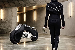 woman wearing black jumpsuit holding gray metal part standing in front of black and gray sports bike