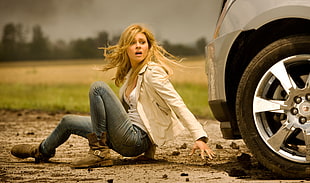 woman wearing beige jacket and gray jeans sitting on ground near vehicle