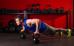 man wearing blue tank top and brown shorts using two black kettle bells