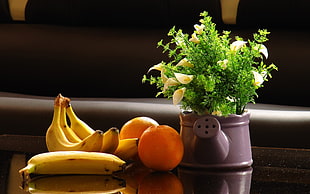 four bananas and two oranges next to watering can pot placed on brown wooden table