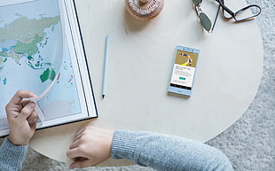 person wearing gray sweater sitting in front table holding world map print beside smartphone