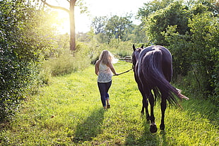 woman in white top and blue denim jeans walking with black horse on green grass during daytime