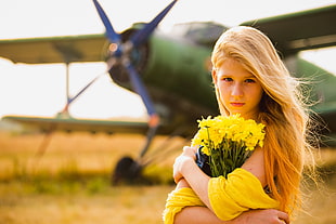 selective focus photography of blonde girl holding yellow flowers against green monoplane