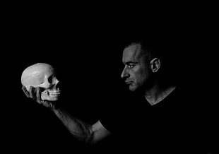 man in v-neck shirt standing holding skull in grayscale photography