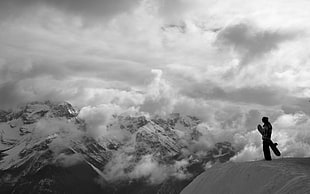 grayscale photo of a person standing on cliff carrying snowboard near snow covered mountain surrounded by clouds HD wallpaper