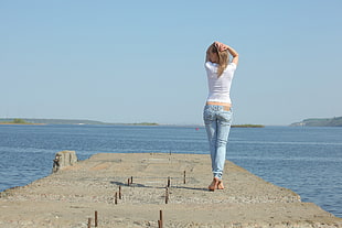 woman in white top and blue denim pants standing on concrete dock over body of water HD wallpaper