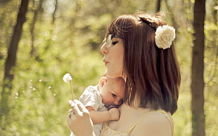 woman carrying baby and holding dandelion flower beside tree