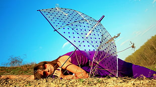 woman wearing purple dress holding transparent umbrella laying in the ground during blue sky