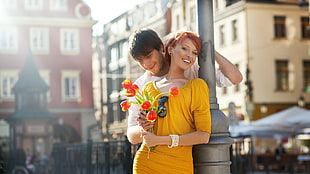 woman in yellow elbow sleeve dress holding tulips leaning at a grey metal post beside a man in white shirt