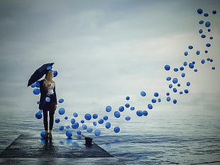 woman with black umbrella standing on dock with blue balloons floating nearby