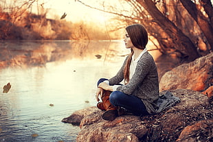 woman wearing blue denim pants and gray cardigan sitting on rock beside river