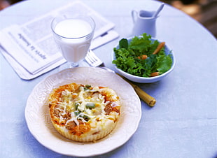 round melted cheese topped food on white ceramic plate beside bowl of green salad and footed glass of milk