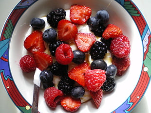 sliced strawberries and blueberries on white, red, and blue ceramic bowl with cream
