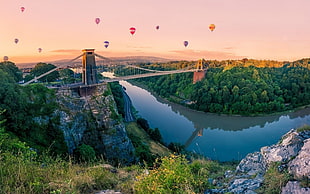 cable bridge surrounded by green trees, water, forest, mountains, hot air balloons