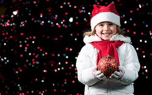 girl wearing white jacket and red christmas hat holding bauble with bokeh background