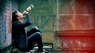 man in blue full-zip hoodie drinking a bottle of liquor while sitting beside wall