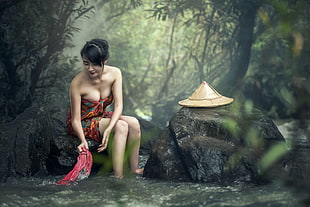 woman wearing black, brown, and green floral towel while sitting on rock washing clothes in the river