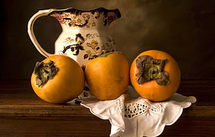 three orange persimmons on brown wooden table in front of white ceramic pitcher