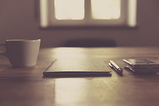 selective photography of black notebook, pen, ceramic mug, and android smartphone on table top