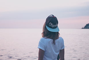 woman in white scoop-neck T-shirt and black-and-teal fitted rim cap outfit standing near sea during daytime