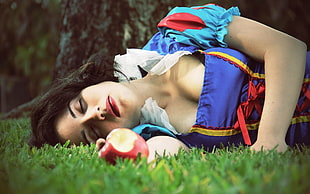 woman in blue and red Snow White costume lying on green open field holding Apple fruit