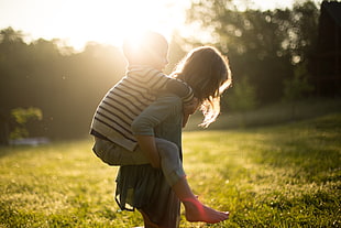 girl lifting the boy on her back on green grass field under sunlight