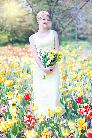 woman in yellow tank dress holding yellow petaled flowers standing on yellow and red petaled flowers field during daytime HD wallpaper
