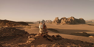 person wearing orange and white suit sitting on brown rocks in distance brown field and rock formation during daytime
