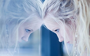woman with blonde hair leaning her head on a glass panel looking to her own reflection