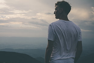 man wearing white t-shirt overlooking black field with hills under cloudy sky during daytime