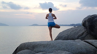 person wearing white tank top and blue shorts while meditating on top of gray stone