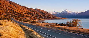 photo of gray asphalt road surrounded with brown mountains near body of water during day time, mt cook