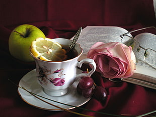 white and pink floral ceramic teacup filled with tea with sliced lemon and teaspoon on white ceramic saucer beside green apple, pink rose flower, eyeglasses and book