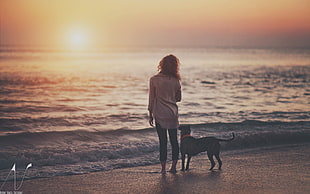 brown haired woman wearing white long sleeve shirt and black pants beside dog standing in seashore