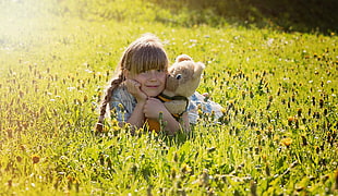 girl with beige and brown animal plush toy laying on green grass