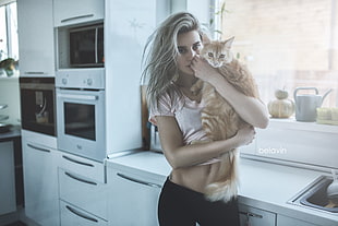 woman wearing white crop top and black bottoms carrying orange tabby cat inside room