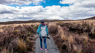 woman in teal jacket, blue jeans, pink sneakers and gray backpack walking in pathway beside brown grasses under blue and white clouds