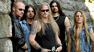 five men wearing black tank top and blue denim vest standing near gray wall during daytime