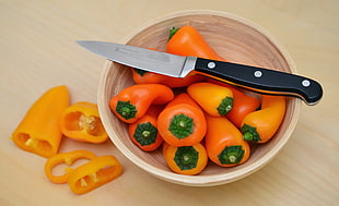 food photography of black handle kitchen knife on top of round brown bowl with a bowl of yellow bell peppers
