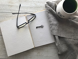 black framed eyeglasses beside black key and white ceramic mug on top of gray textile and white book