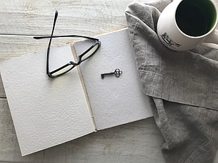 black framed eyeglasses beside black key and white ceramic mug on top of gray textile and white book