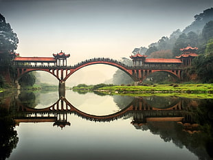 brown bridge mirrored on calm body of water with green grass field under white sky during daytime