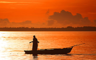 silhouette photo of person holding fish net standing on canoe on body of water under orange sky