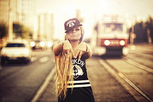 shallow focus photography of woman in black and white tank top and black and white snapback cap in middle of road with tram and cars